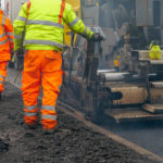 Two construction workers, in high visibility workwear, walking beside an asphalt paver machine which is laying new tarmac on a public road. Using a control panel one of the workmen is controlling the distribution of hot, steaming asphalt concrete, making sure the level of tarmac is consistent. It is slightly compacted, before an asphalt drum compactor follows to roll it further before the new tarmac sets.
